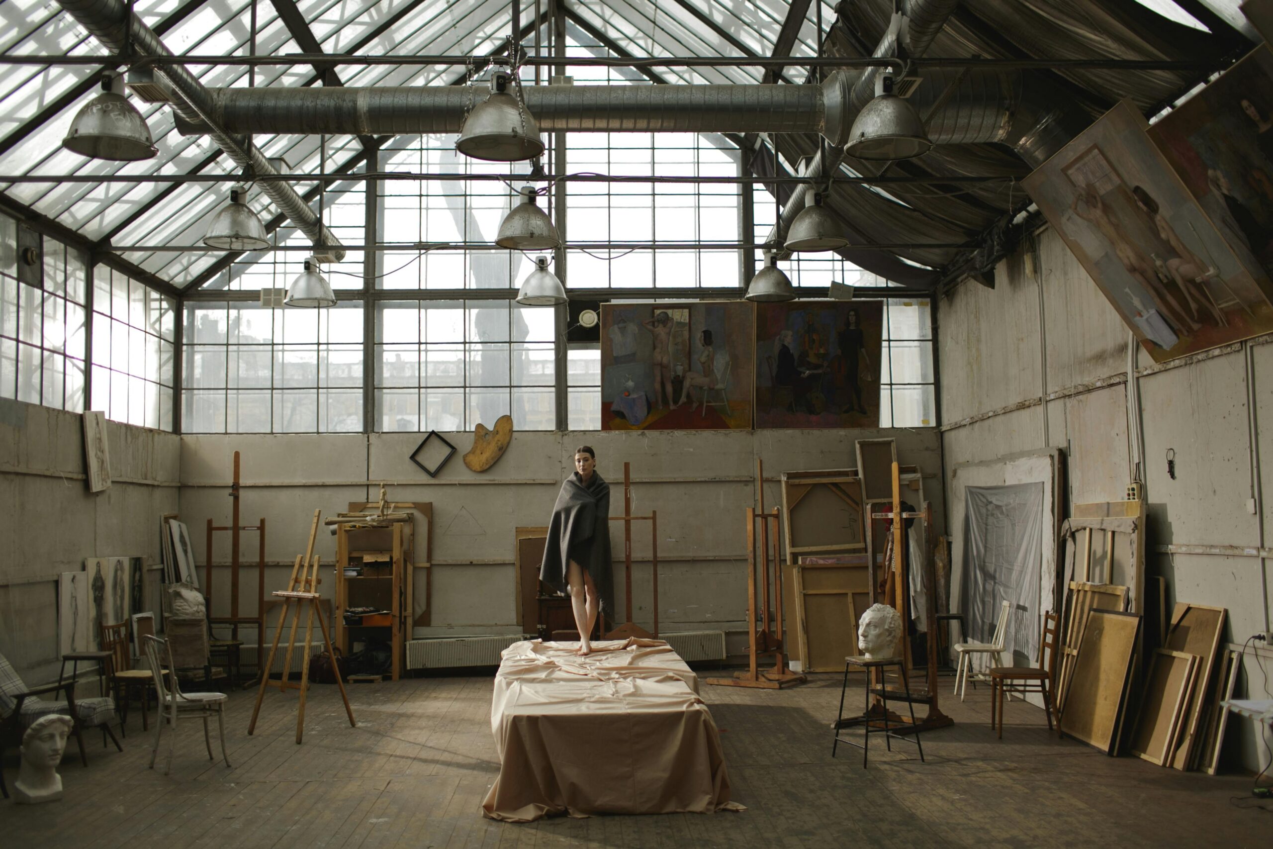 A model stands on a table amidst artistic clutter in a spacious, sunlit studio.